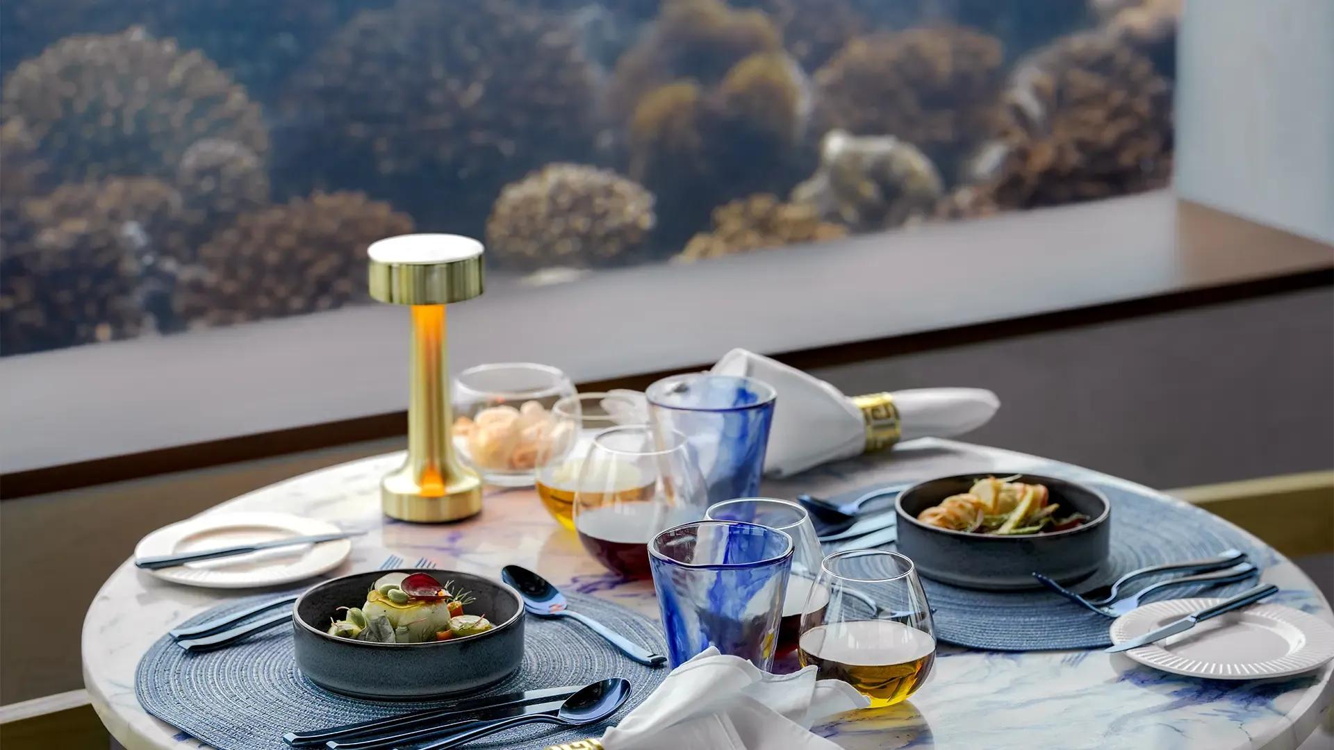 Стол в Only BLU с блюдами и бокалами у стекла, за которым видны кораллы, OBLU Select Lobigili