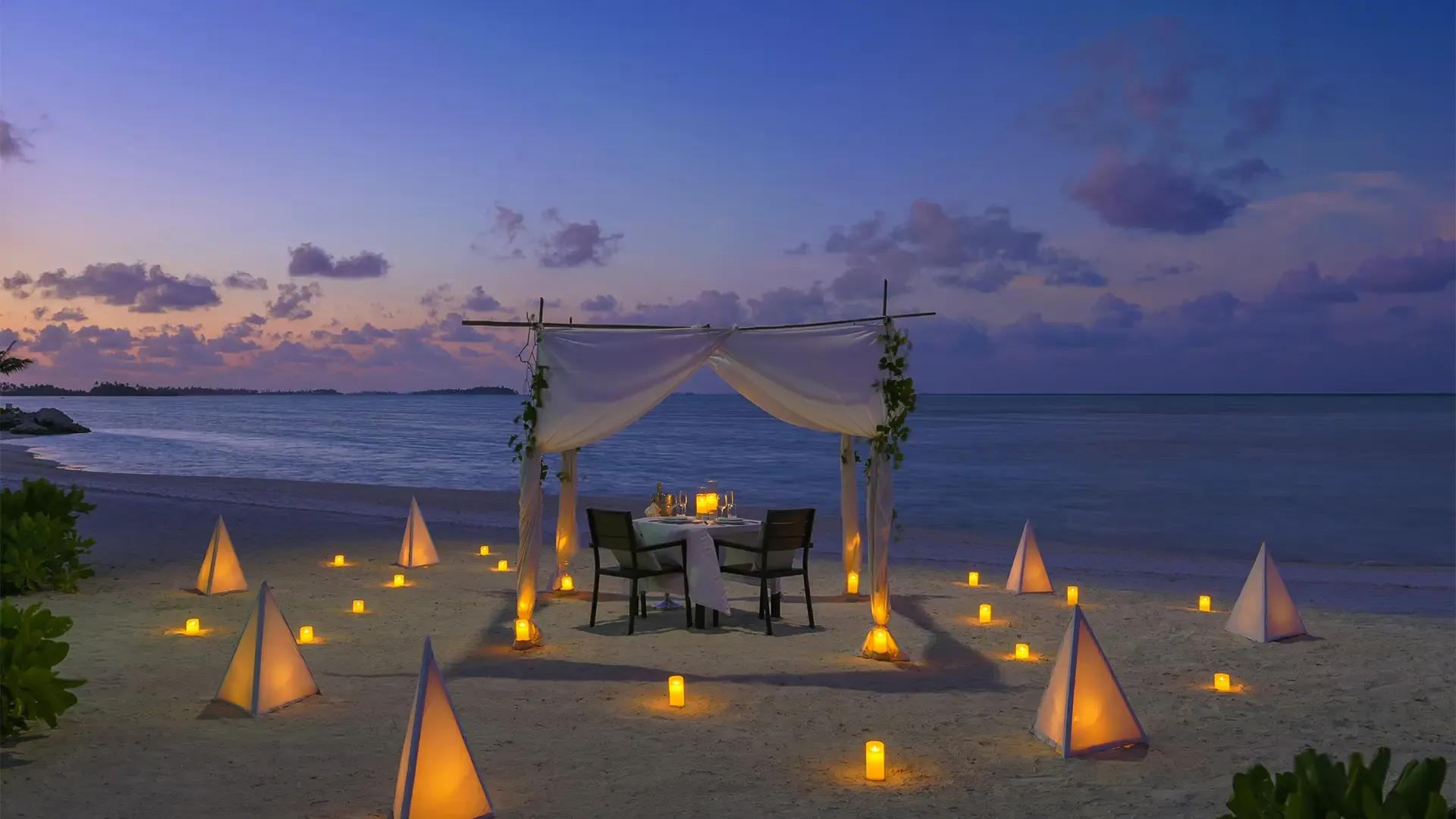 Беседка с белой тканью и накрытый стол для ужина на пляже Rahaa Resort на закате