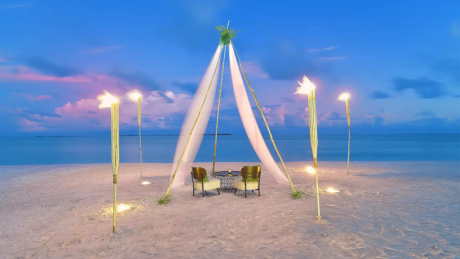 Шатёр из белой ткани и два кресла на пляже Rahaa Resort в сумерках у воды